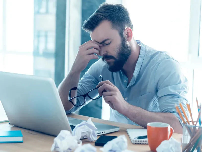 Stressed man rubbing eyes at his desk with glasses in hand, showing symptoms of eye fatigue and dry eye