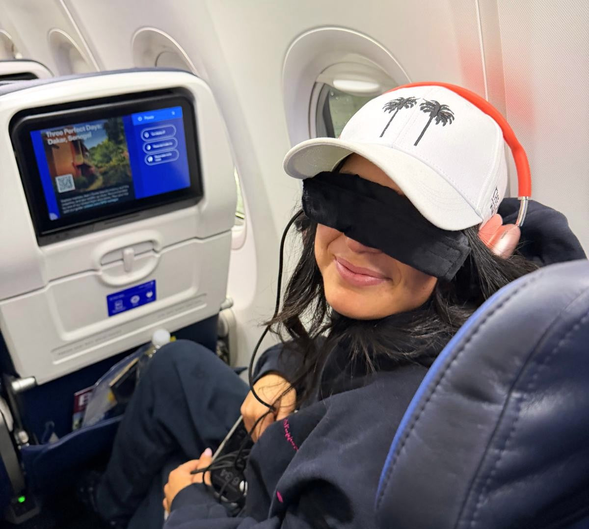 Person sitting in an airplane seat with a heated dry eye mask on, wearing a cap with palm tree designs.